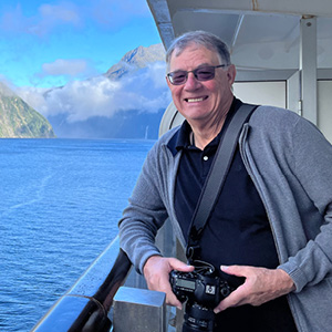 A man standing on a boat, smiling and holding a camera, with a body of water and mountains visible behind him