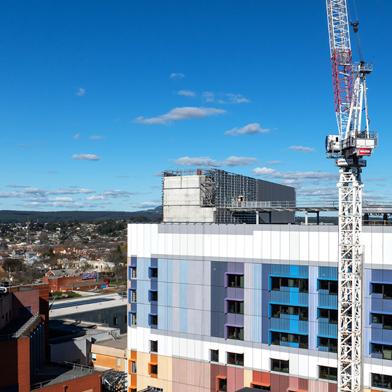 New multi-storey hospital building under construction with colourful exterior panels, rooftop scaffolding and a tower crane, overlooking the Ballarat city skyline under a clear blue sky.