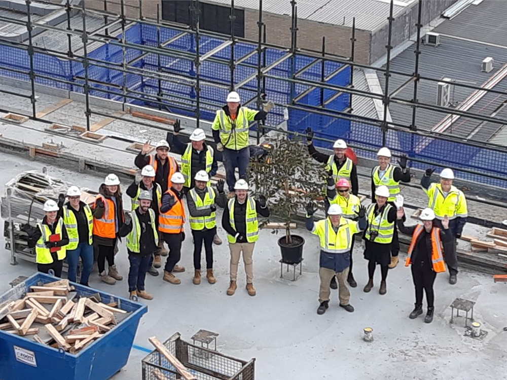 A group of construction workers wearing construction vests and hates and smiling and waving.