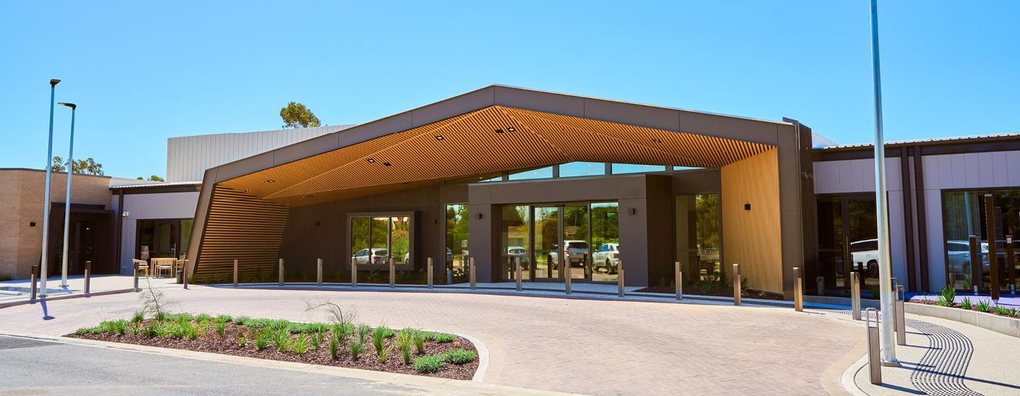 A modern building with a large overhanging entrance and a glass facade. The area features a roundabout driveway with landscaped greenery. 