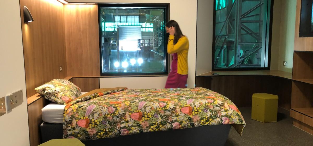 inside a purpose-built, full-scale bedroom prototype, a woman stand next to a bed with a floral bedcover