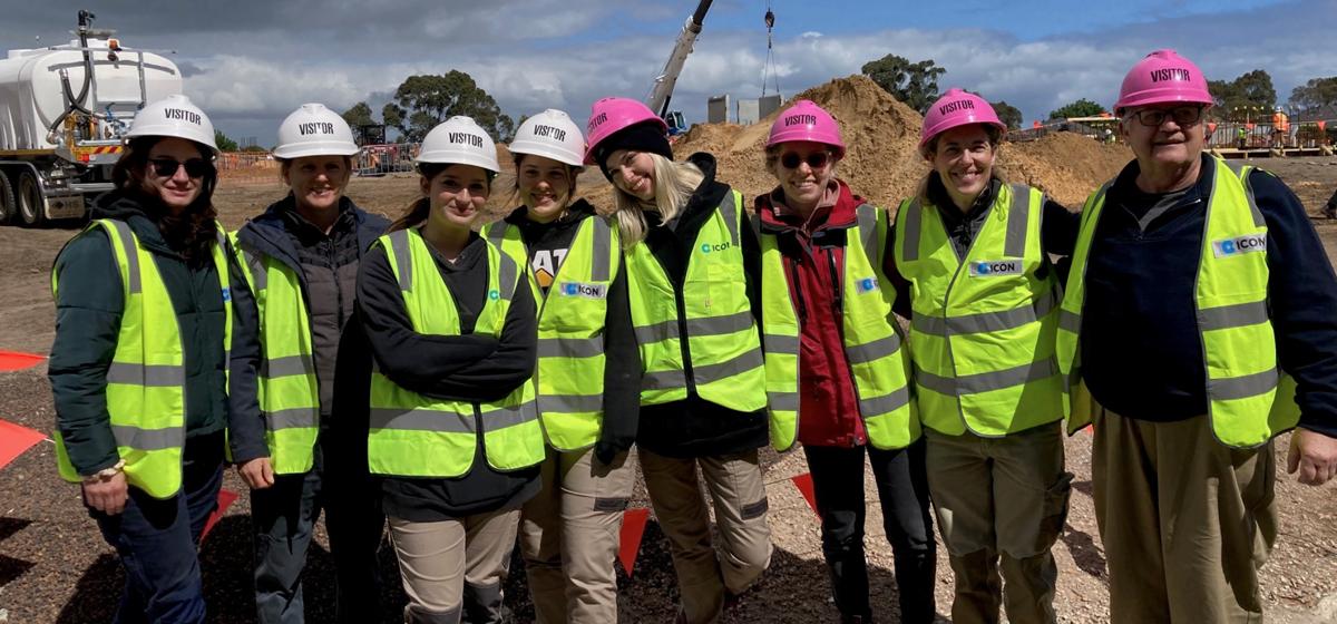 A group of women standing at a construction site and smiling wearing white and pink hard hats and hi-vis vests