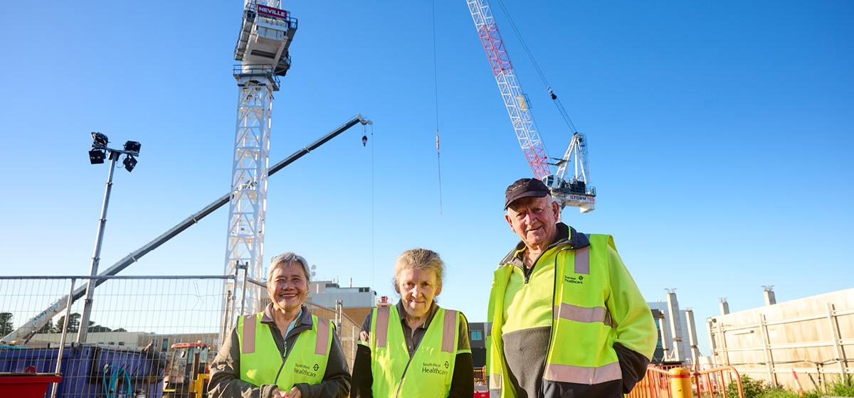Three community members wearing high-visibility clothing standing in front of a hospital construction site with cranes in the background