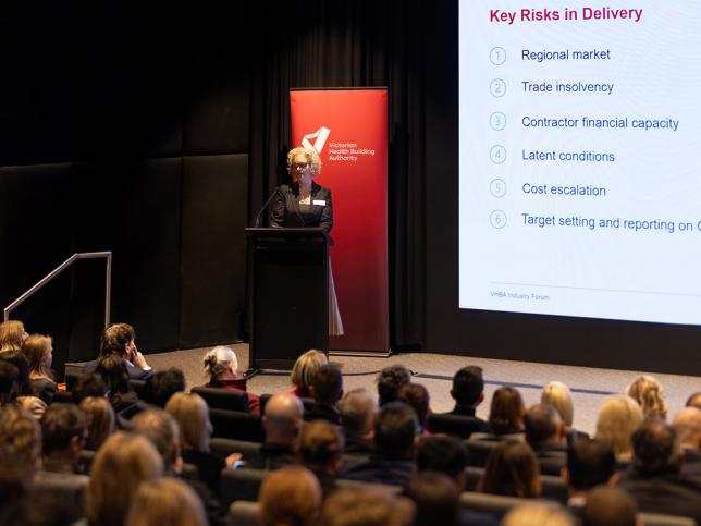 Jodie Gervasoni, COO at the Victorian Health Building Authority, stands at a lectern on a stage and gestures at a graph projected onto a screen beside her