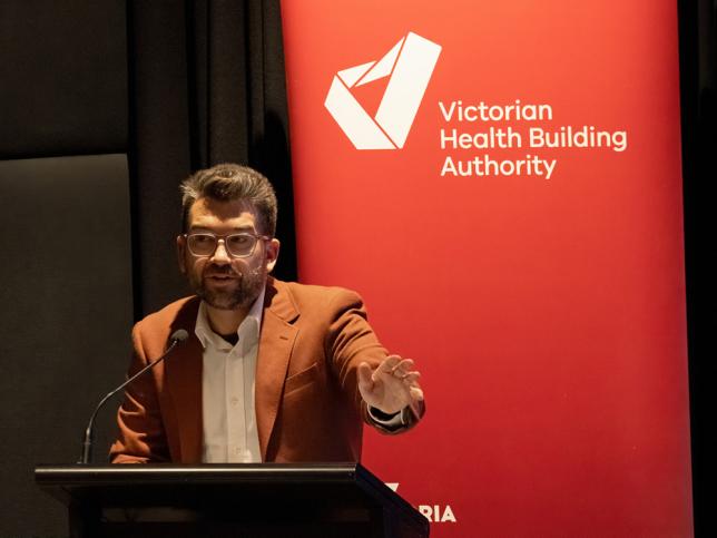 The Honourable Josh Bull, Parliamentary Secretary for Infrastructure Delivery, stands at a lectern delivering a speech. The VHBA logo is visible on a banner behind him