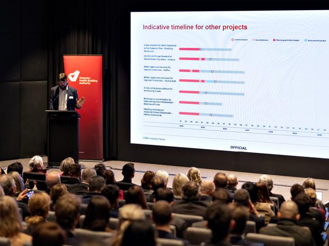 Trent Burke, Senior Executive Director at the Victorian Health Building Authority, stands at a lectern on a stage and gestures at a graph projected onto a screen beside him