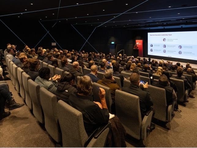 People sitting in seats in a lecture hall, looking towards a stage with a projection lit on the wall behind it