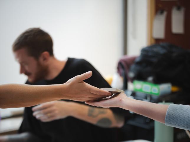 A clay piece is handed over from one person's hand to another person's hand, with Dan Elborne in the background