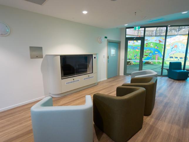 Interior of a modern hospital lounge area with comfortable chairs, a large TV, and a vibrant mural visible through the window.