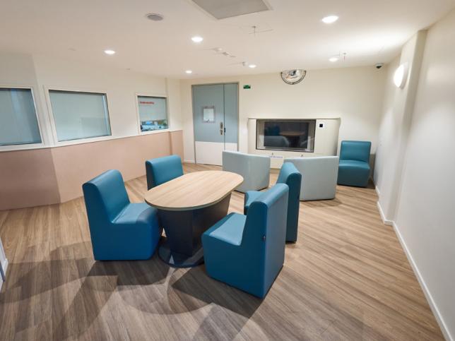 Interior of a modern lounge featuring a round wooden table with blue chairs, adjacent to a seating area with couches and a wall-mounted television.