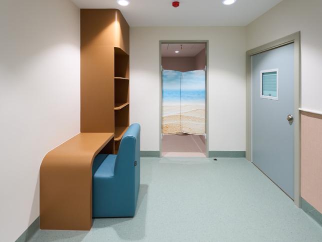 Interior of a modern medical waiting room featuring a curved wooden desk and bookshelf, a blue chair, and doors with a beach-themed mural.