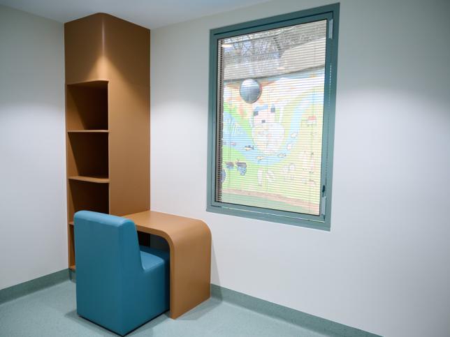 Interior of a small room with an empty bookshelf, a blue chair, and a window showing a colorful mural outside.