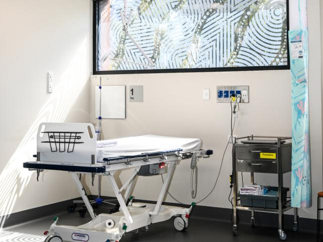 Medical treatment room with an adjustable bed, medical equipment cart, and art on the window.