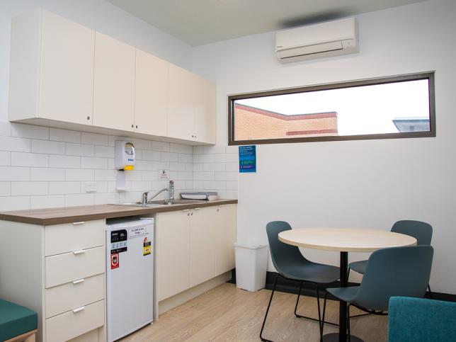 Modern kitchenette and adjacent small dining area with a round table and teal chairs, equipped with cabinets, a sink and under-cabinet refrigerator.