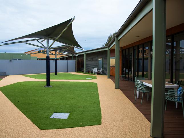 Outdoor courtyard area with paths, a sail shade, synthetic grass, and a table and chairs undercover.