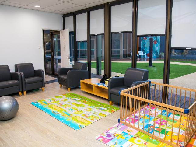 Interior of a bright children's playroom with sofas, colourful floor mats, a playpen and large windows show an outdoor view.