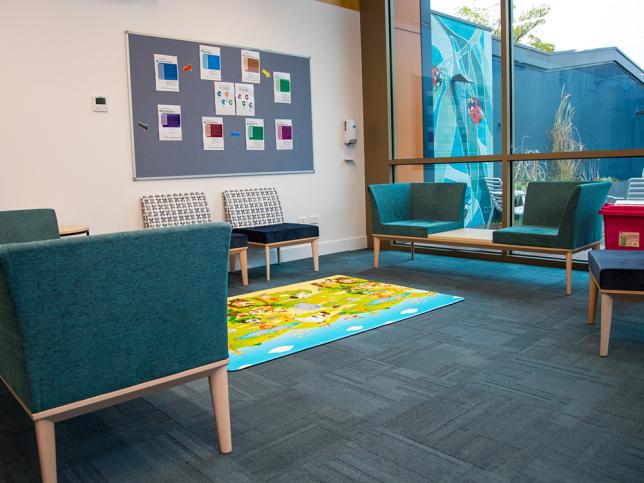 A modern waiting room with colourful chairs, a children's play mat, and a bulletin board alongside a large window.