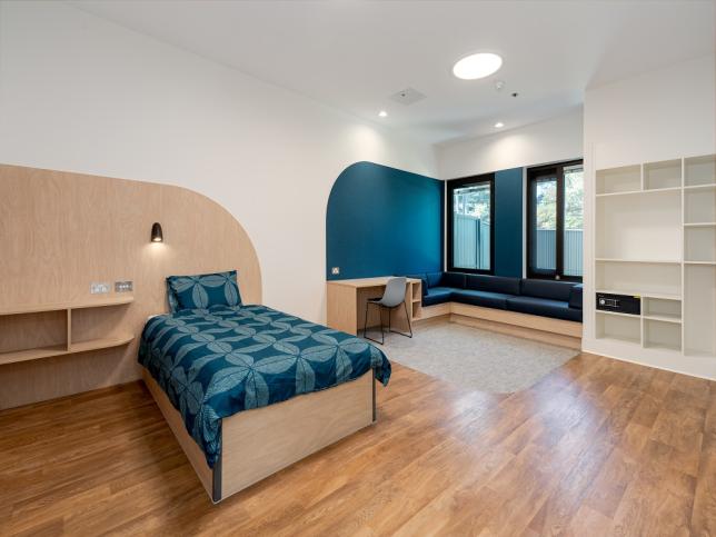 A modern, spacious room featuring a single bed with a blue patterned bedspread, a built-in light, and a small wooden desk with a chair. 
