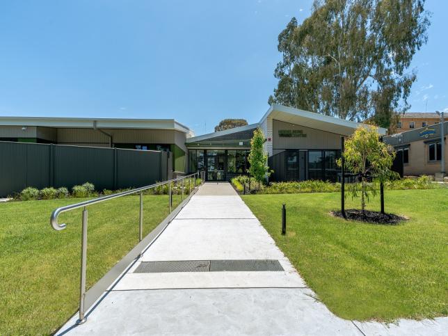 The exterior of a modern building surrounded by well-maintained green lawns and trees. A wide, paved pathway with metal railings leads to the entrance.