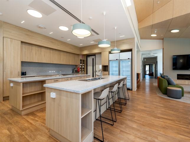 A modern kitchen features light wooden cabinets, a spacious island with four stools and contemporary hanging lights. To the right, a living area has a wall-mounted TV and a green sofa.