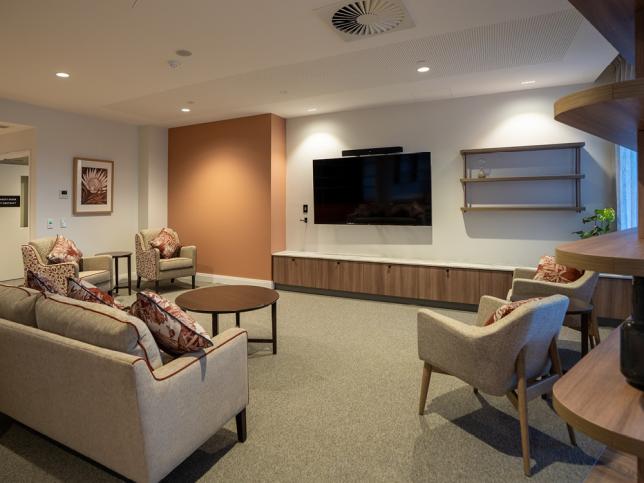 Comfortable shared lounge in a residential aged care home, featuring sofas, armchairs, coffee tables and a wall-mounted television in a warm, modern setting.