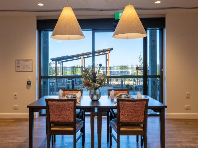 Small dining area in a residential aged care home with a set table, pendant lighting and large windows overlooking an outdoor courtyard.