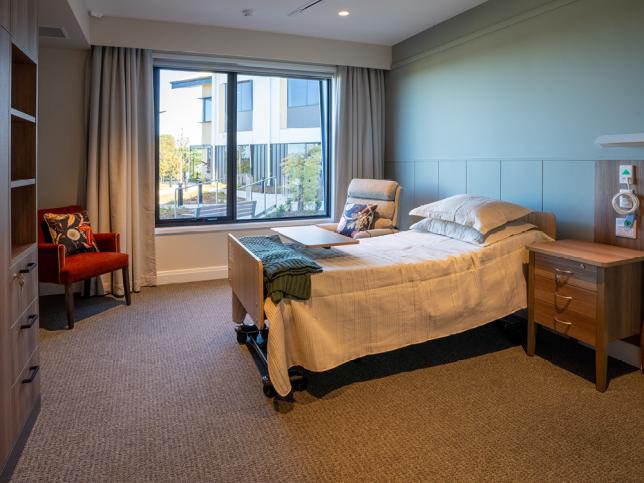 Private aged care resident room with an adjustable bed, bedside table, armchair and large window providing natural light and views of landscaped grounds.