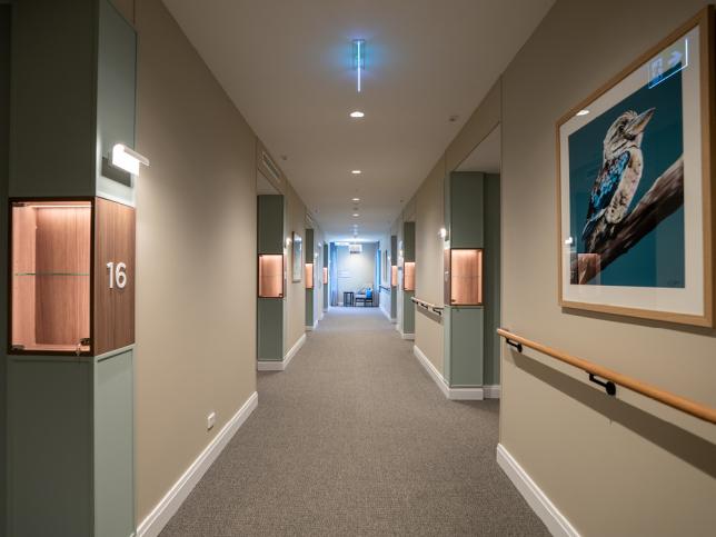 Wide, carpeted corridor in a residential aged care facility with handrails, room entrances and framed artwork along the walls.