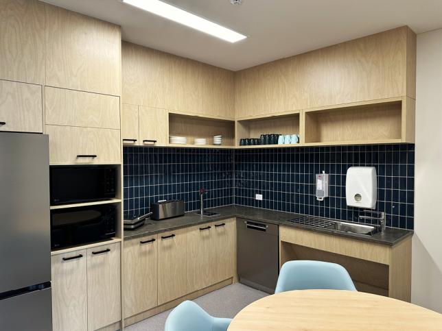 Staff kitchen with light timber cupboards, dark blue tiles, sinks and a round table.