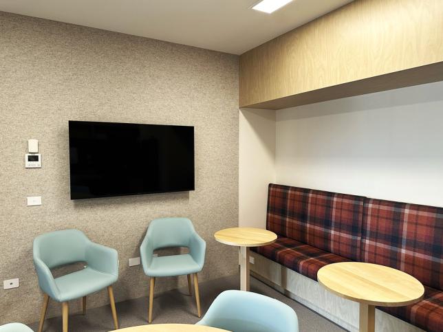 Staff lounge with pale blue chairs, plaid bench seating, tables and a wall-mounted screen.