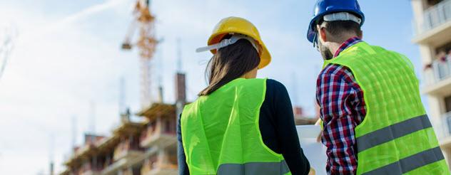 Image of a female and male workers reviewing a document together on a construction site. They are dressed in high vis vests and hard hats, and facing away from the camera.