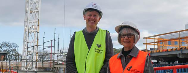 a man and a woman in hi-vis vests and helmets stand smiling infront of cranes