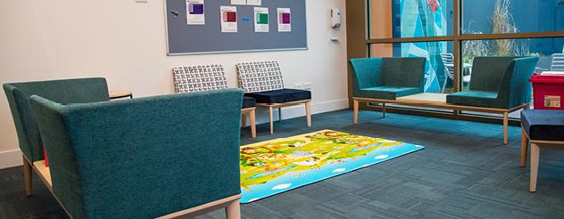 Interior of a waiting area with teal and patterned chairs, small wooden tables, colourful rug, and bulletin board on the wall.