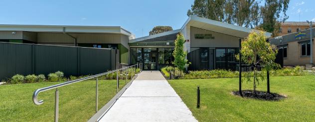 The exterior of a modern building surrounded by well-maintained green lawns and trees. A wide, paved pathway with metal railings leads to the entrance.