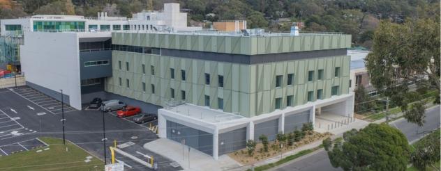 Aerial view of the new Angliss Hospital building. It is a modern multi-storey hospital with a sage green and grey patterned facade. A car park, covered entrance and young trees sit in the foreground, with suburban streets and bushland behind.