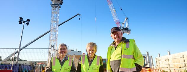 Three people wearing high-visibility vests stand at a construction site, with cranes and building works visible in the background.