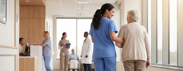 A medical professional and elderly woman walking along a corridor together.